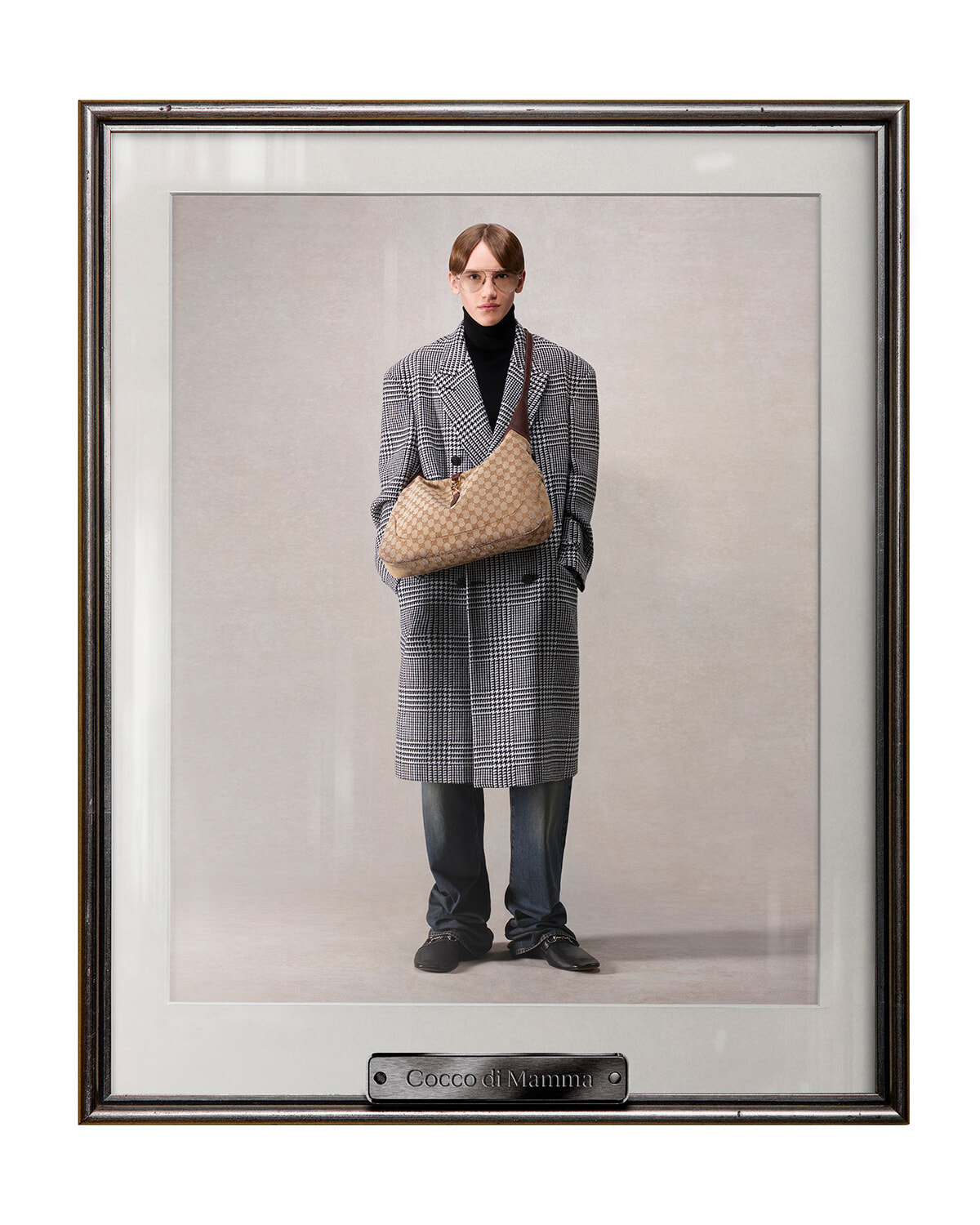 Model wearing a gray plaid overcoat over a black turtleneck, holding a beige patterned shoulder bag in front of a plain backdrop (framed portrait).