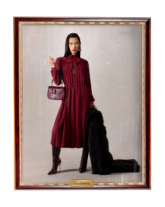 Woman in a burgundy midi dress with a bow at the neck, standing with a fur coat and a matching burgundy handbag and crocodile-embossed boots beside a golden-framed display labeled 'Ereditiera'.