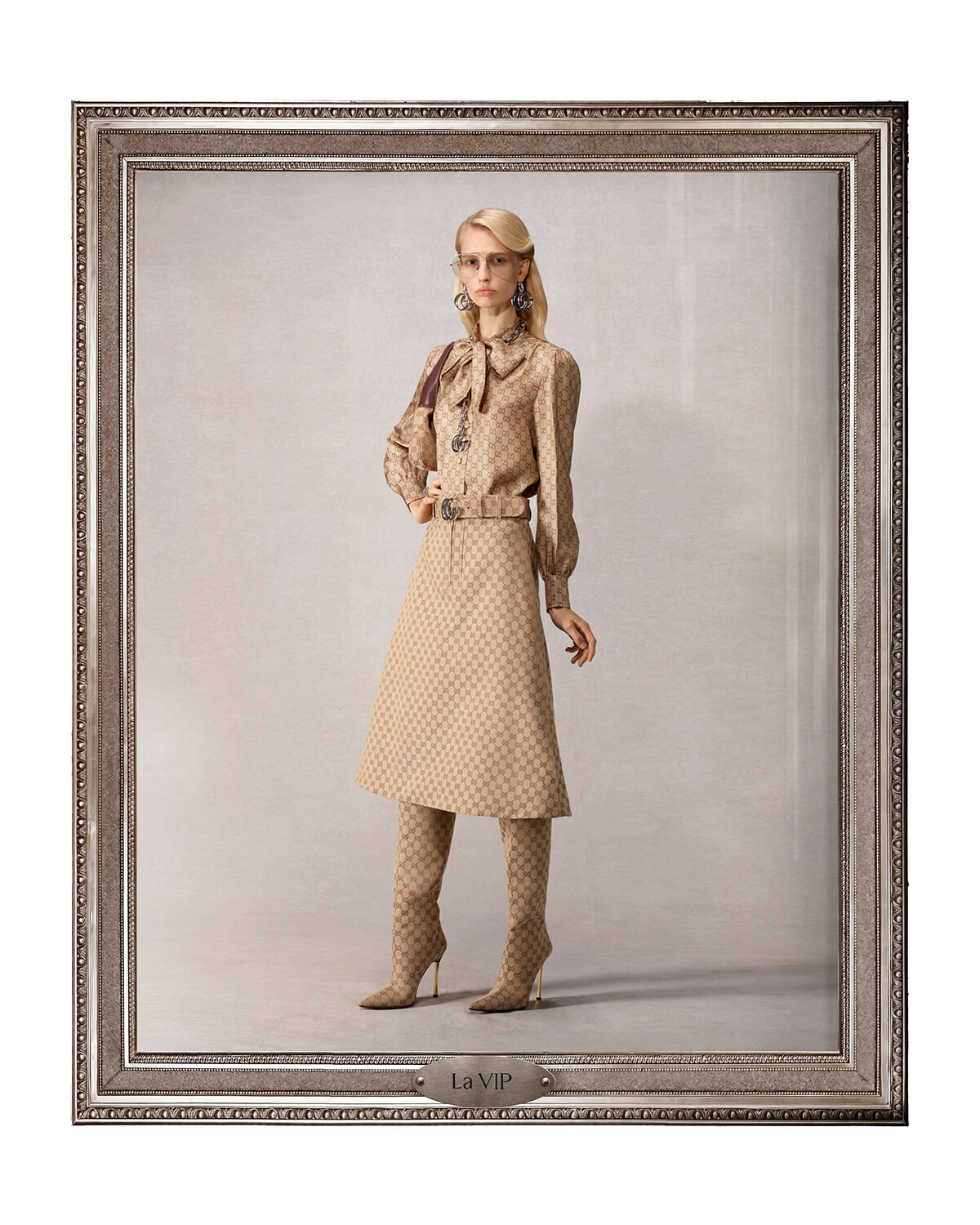 Model in a beige monogram dress with matching knee-high boots, bow blouse, and belt, posed in an ornate gallery frame.