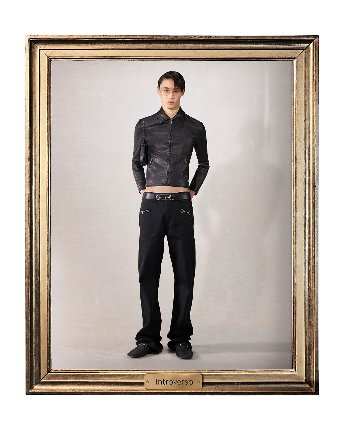 Male model in a black leather jacket and pants, standing with hands in pockets inside an ornate gold-framed portrait frame that reads 'Introverso' at the bottom plaque.