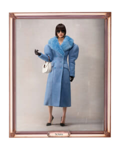 Woman modeling a long blue coat with a fluffy blue fur collar, black gloves, oversized sunglasses, and a white handbag, standing in a neutral studio setting framed like a portrait labeled 'Sciura'