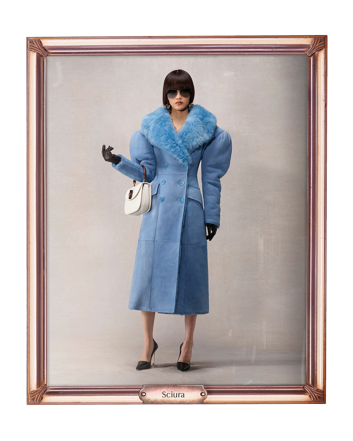 Woman modeling a long blue coat with a fluffy blue fur collar, black gloves, oversized sunglasses, and a white handbag, standing in a neutral studio setting framed like a portrait labeled 'Sciura'