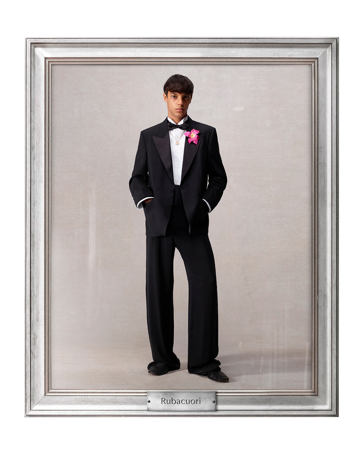 Young man in a black tuxedo with bow tie and pink boutonniere stands with hands in pockets, framed as a formal portrait labeled Rubacuori.