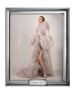 Portrait-style fashion photo of a model wearing a long pale pink fluffy fur coat, standing in a studio with a neutral backdrop, inside a silver ornate frame labeled 'La Drama Queen'.
