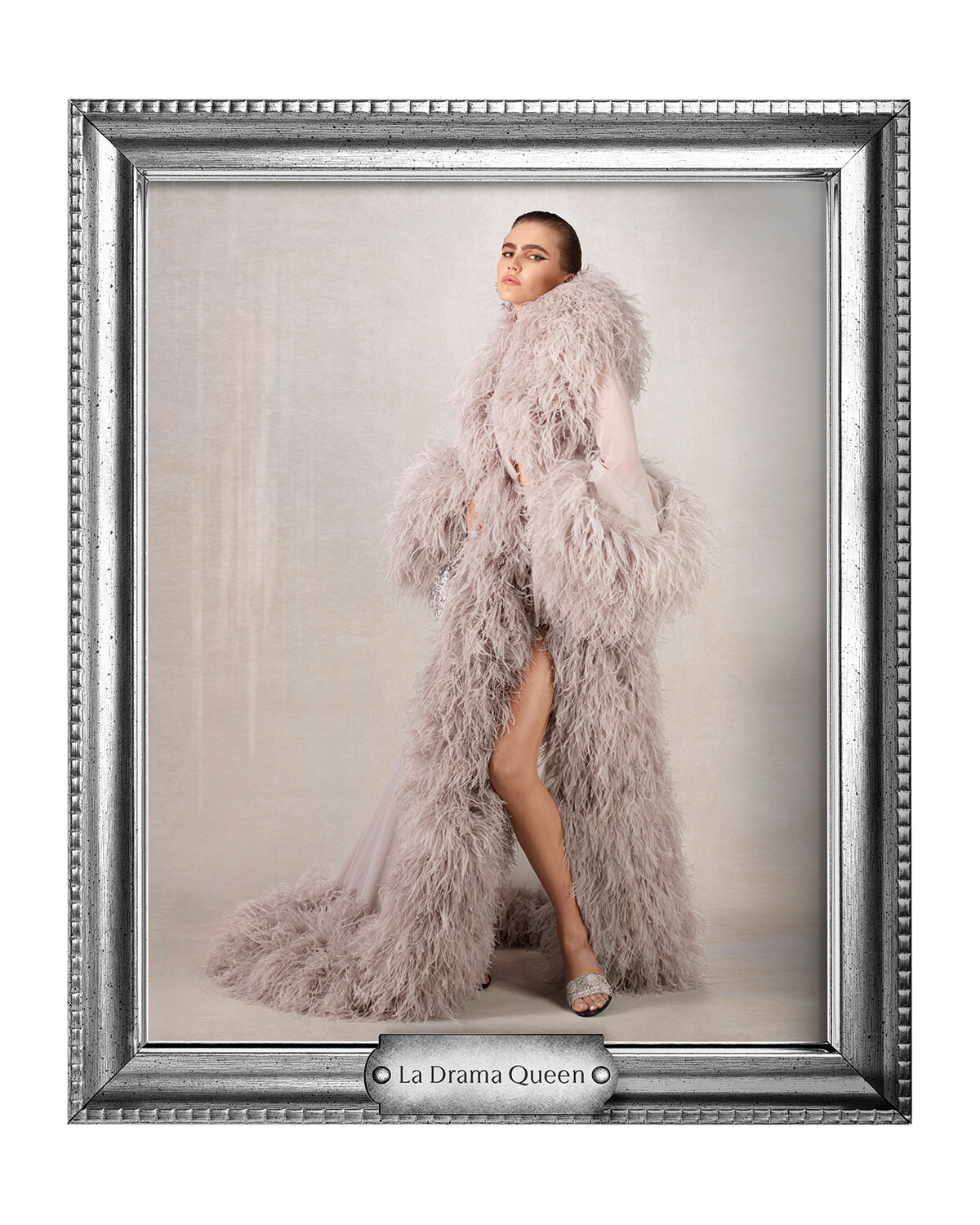 Portrait-style fashion photo of a model wearing a long pale pink fluffy fur coat, standing in a studio with a neutral backdrop, inside a silver ornate frame labeled 'La Drama Queen'.