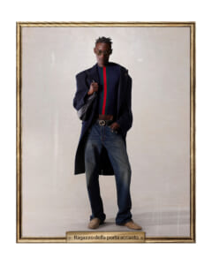 Portrait-style photo of a stylish man wearing a navy long coat, striped knit sweater, jeans, and sunglasses with a shoulder bag.