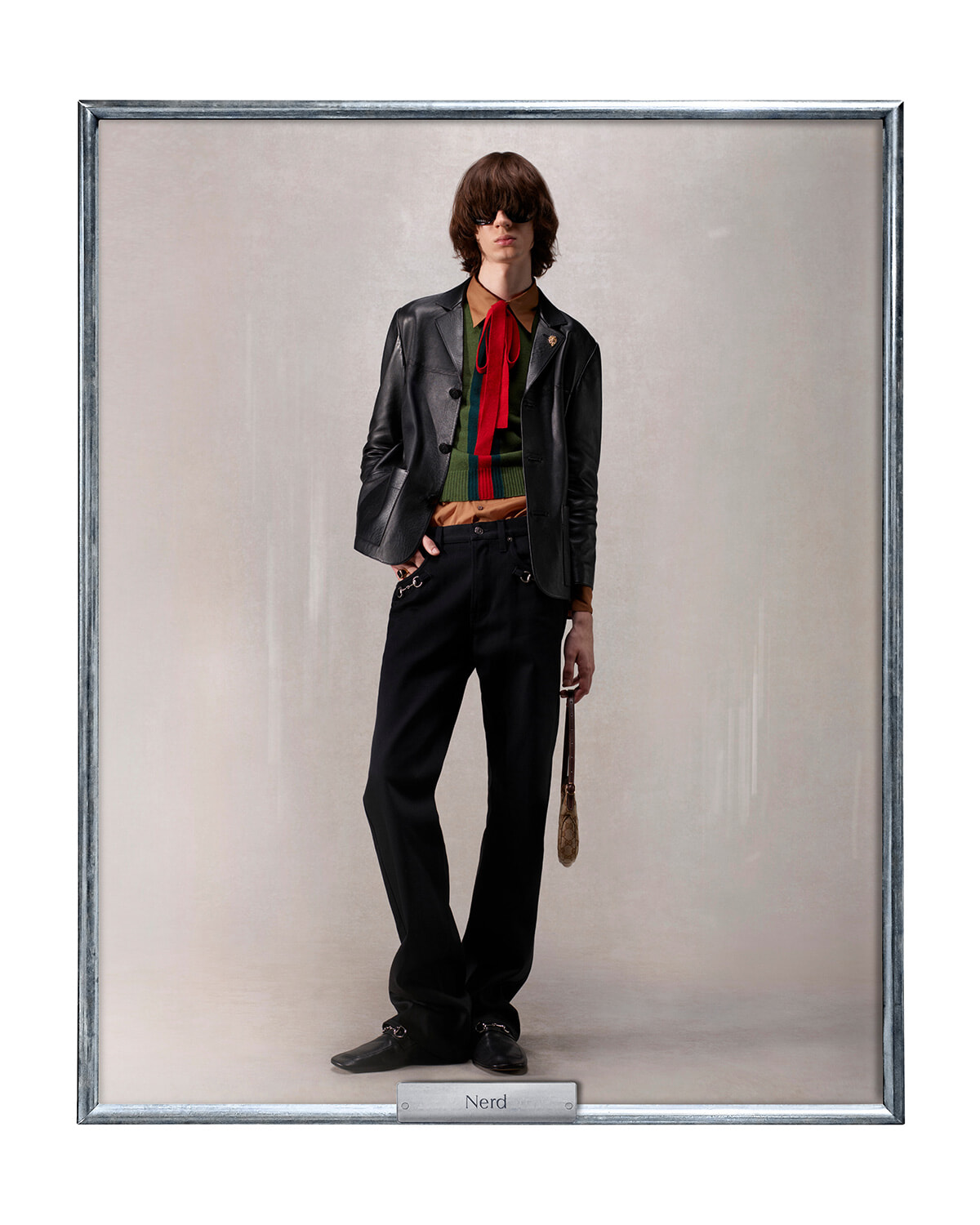 Model in a retro nerd-inspired outfit: black leather blazer over a color-block green sweater with a red tie, brown shirt, black wide-leg pants, sunglasses, and a small bag against a neutral backdrop.