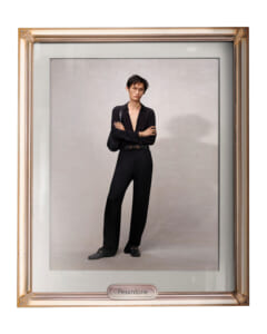 Man in a black outfit with a deep V-neck and belt, arms crossed, posing against a light gray backdrop inside a gold gallery frame labeled Pesantone.