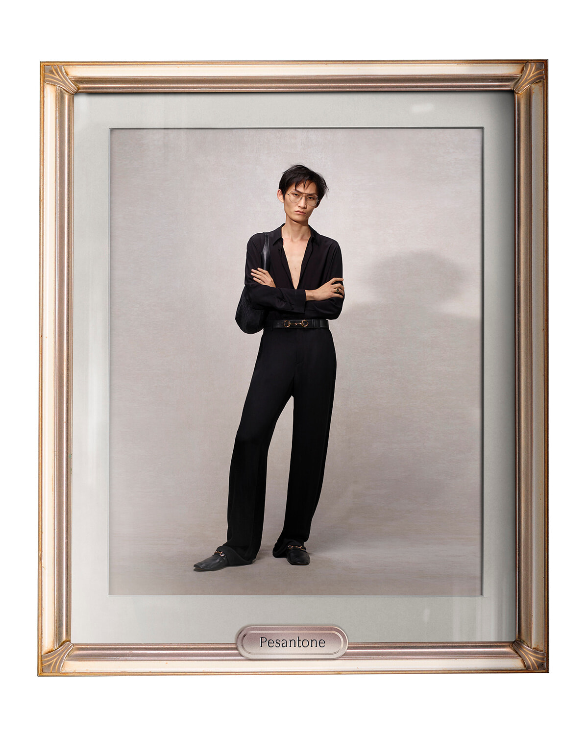 Man in a black outfit with a deep V-neck and belt, arms crossed, posing against a light gray backdrop inside a gold gallery frame labeled Pesantone.