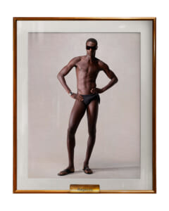 Studio portrait of a tall, muscular Black man in black swim briefs and sunglasses, standing with hands on hips, in a framed display labeled Ragazzo.
