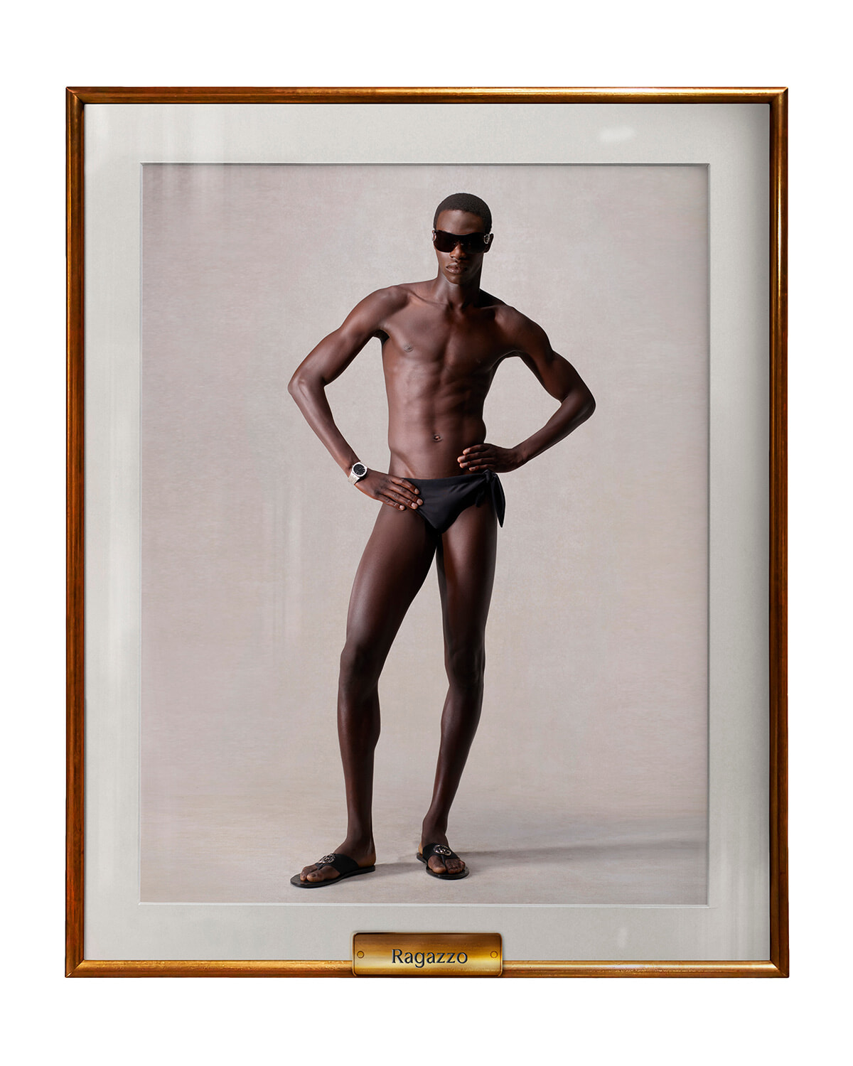 Studio portrait of a tall, muscular Black man in black swim briefs and sunglasses, standing with hands on hips, in a framed display labeled Ragazzo.