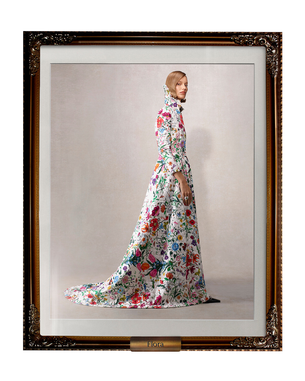 Model wearing a long white floral gown with a high collar, standing in profile inside an ornate frame labeled Flora.