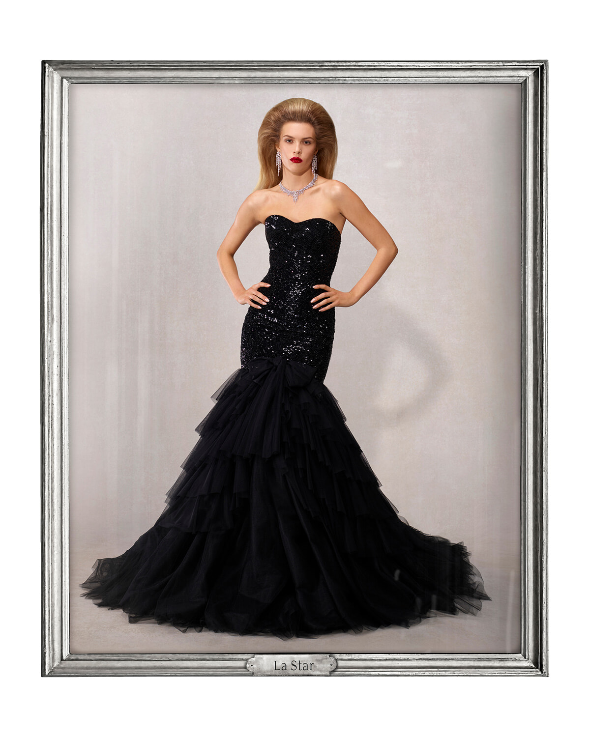 Elegant fashion portrait of a woman in a strapless black sequin gown with a layered tulle mermaid skirt, posing with hands on hips in a silver-framed display.