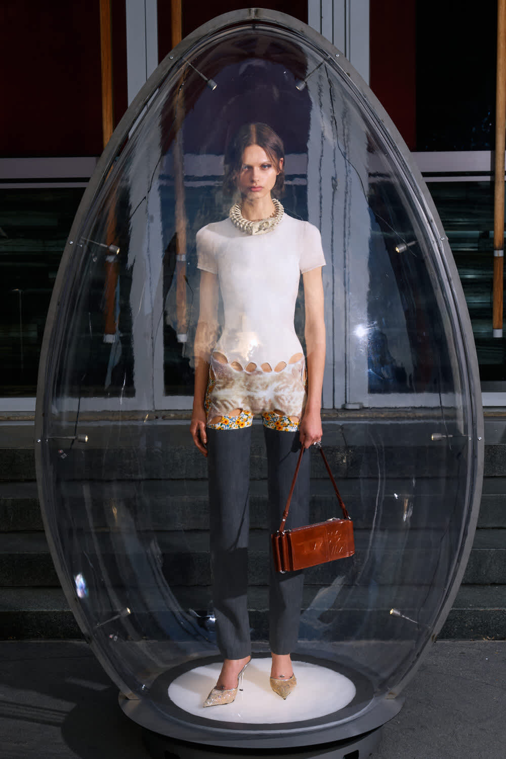 Model stands inside a clear glass capsule on a white circular base, wearing a white top, patterned shorts, dark trousers, gold pointed shoes, and a brown handbag.