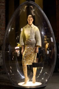 Model in a clear glass capsule wearing a beige coat and floral skirt, holding a black handbag for a fashion exhibit, with brick walls in the background.