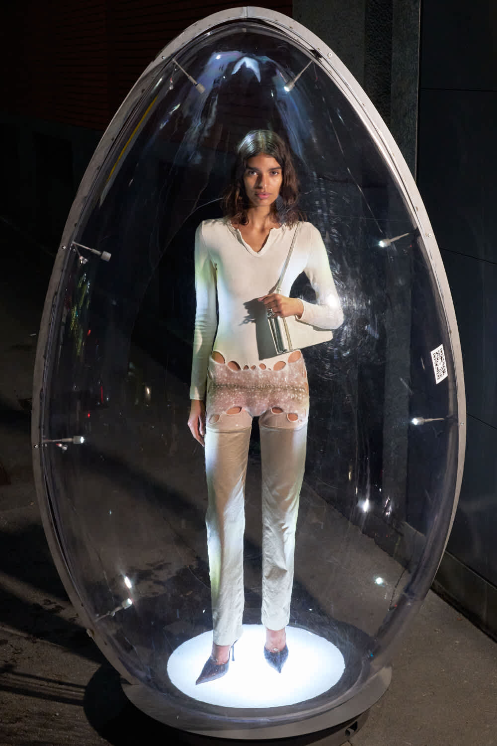 Woman in a pale outfit stands inside a transparent, egg-shaped capsule lit from below in a dim space, holding a bag.
