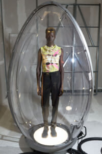 Person wearing a colorful sleeveless top and dark pants stands inside a transparent egg-shaped capsule in a gallery installation, illuminated from below.