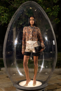 Model stands inside a clear egg-shaped display at night, wearing a sequined brown top, white cut-out shorts, beige heels, and holding a black clutch necklace.