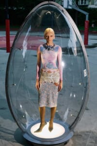 Model inside a clear glass capsule wearing a pastel dress, standing on a lit circular base outdoors.