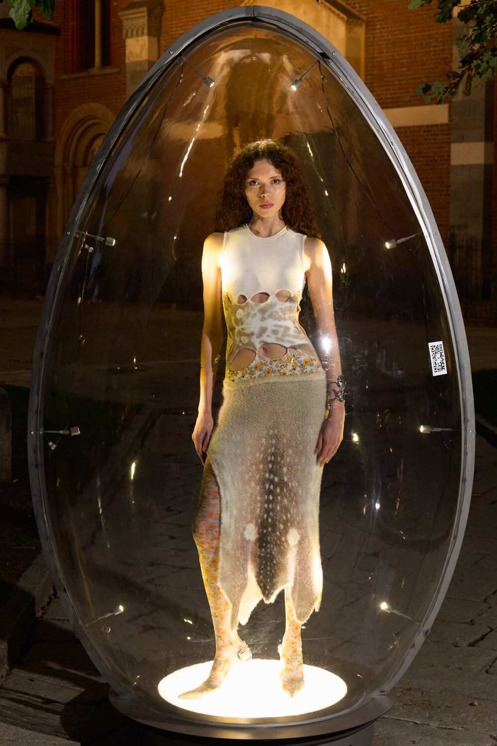 Model stands inside a large glass capsule at night, wearing a sleeveless white dress with cutouts and a long textured skirt, lit from below.