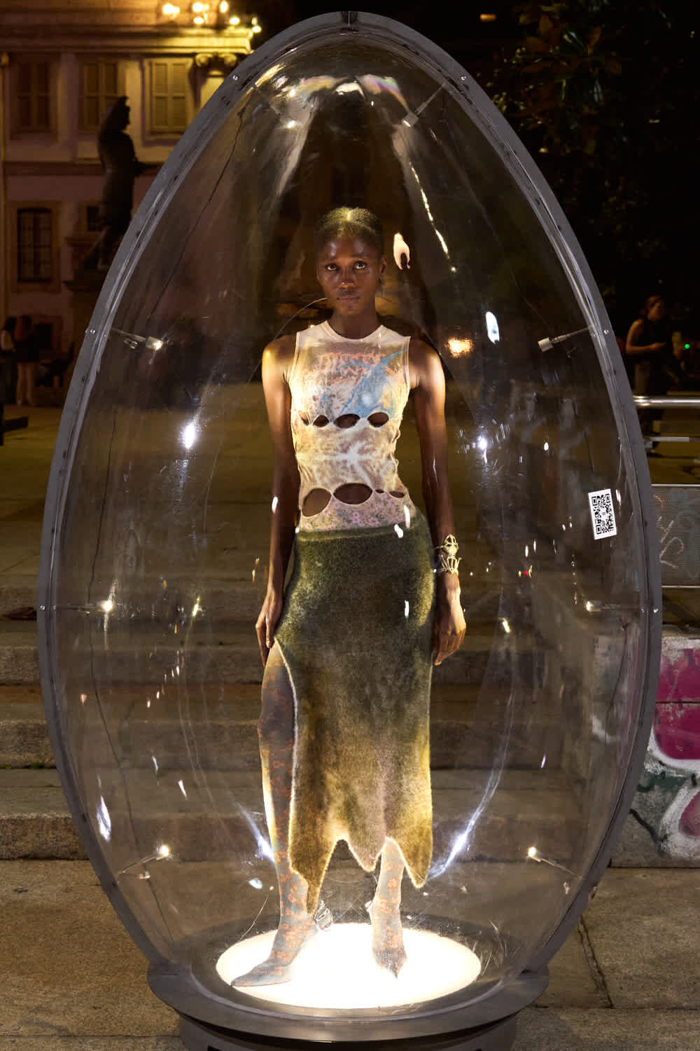 Model wearing a distressed sleeveless top and long textured skirt stands inside a clear glass dome illuminated from below at night.