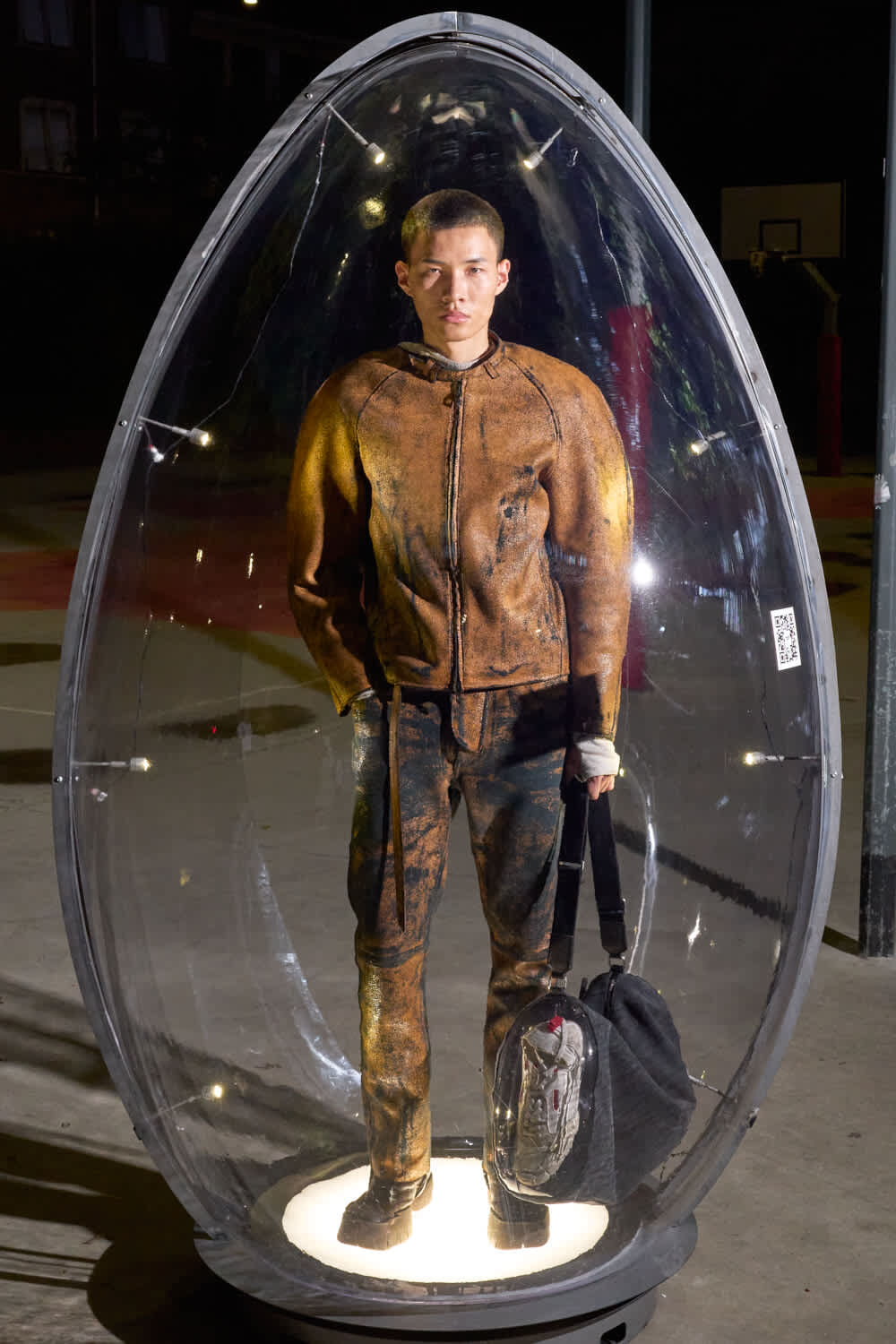 Man wearing a dirty brown jumpsuit standing inside a clear egg-shaped capsule, illuminated from below at night, holding a bag