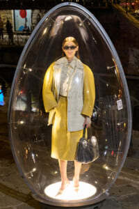 Person in a yellow coat and sunglasses stands inside a transparent glass capsule at night, holding a black handbag.