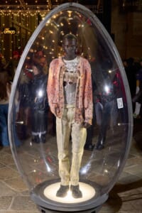 Model inside a clear, egg-shaped display pod at a fashion event, wearing a pink textured cardigan over a graphic tee and beige pants.