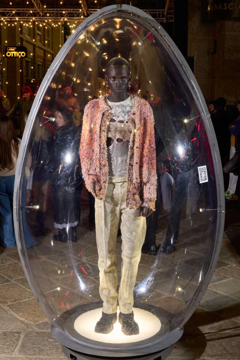 Model inside a clear, egg-shaped display pod at a fashion event, wearing a pink textured cardigan over a graphic tee and beige pants.