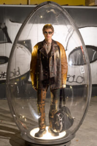 A life-sized mannequin wearing a brown-to-black leather coat and sunglasses inside a clear glass capsule, on display in a graffiti-covered setting.