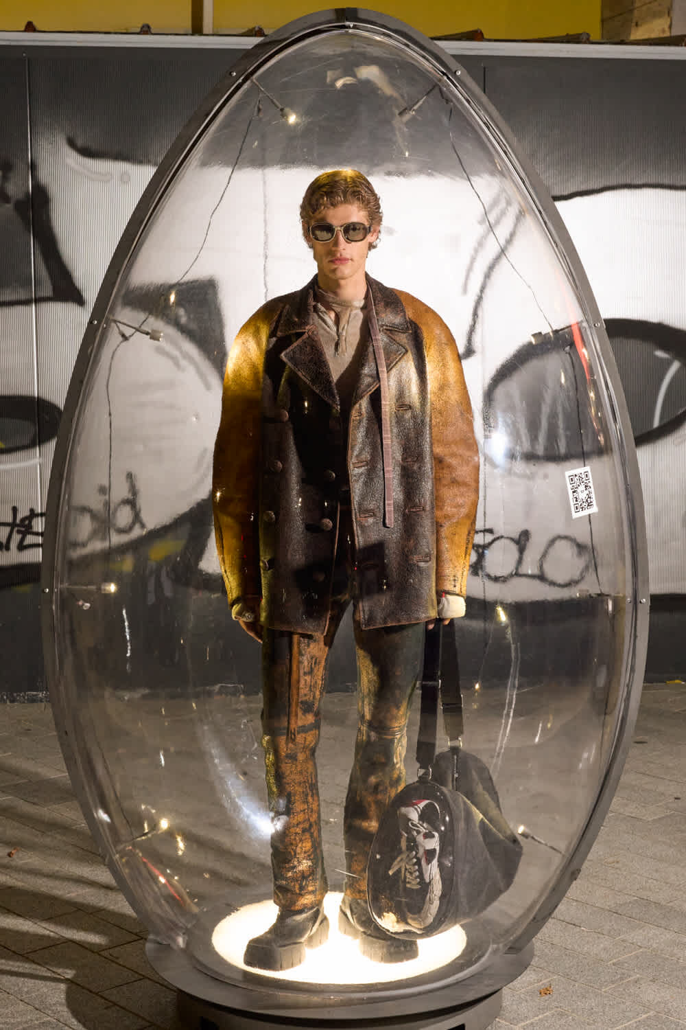 A life-sized mannequin wearing a brown-to-black leather coat and sunglasses inside a clear glass capsule, on display in a graffiti-covered setting.