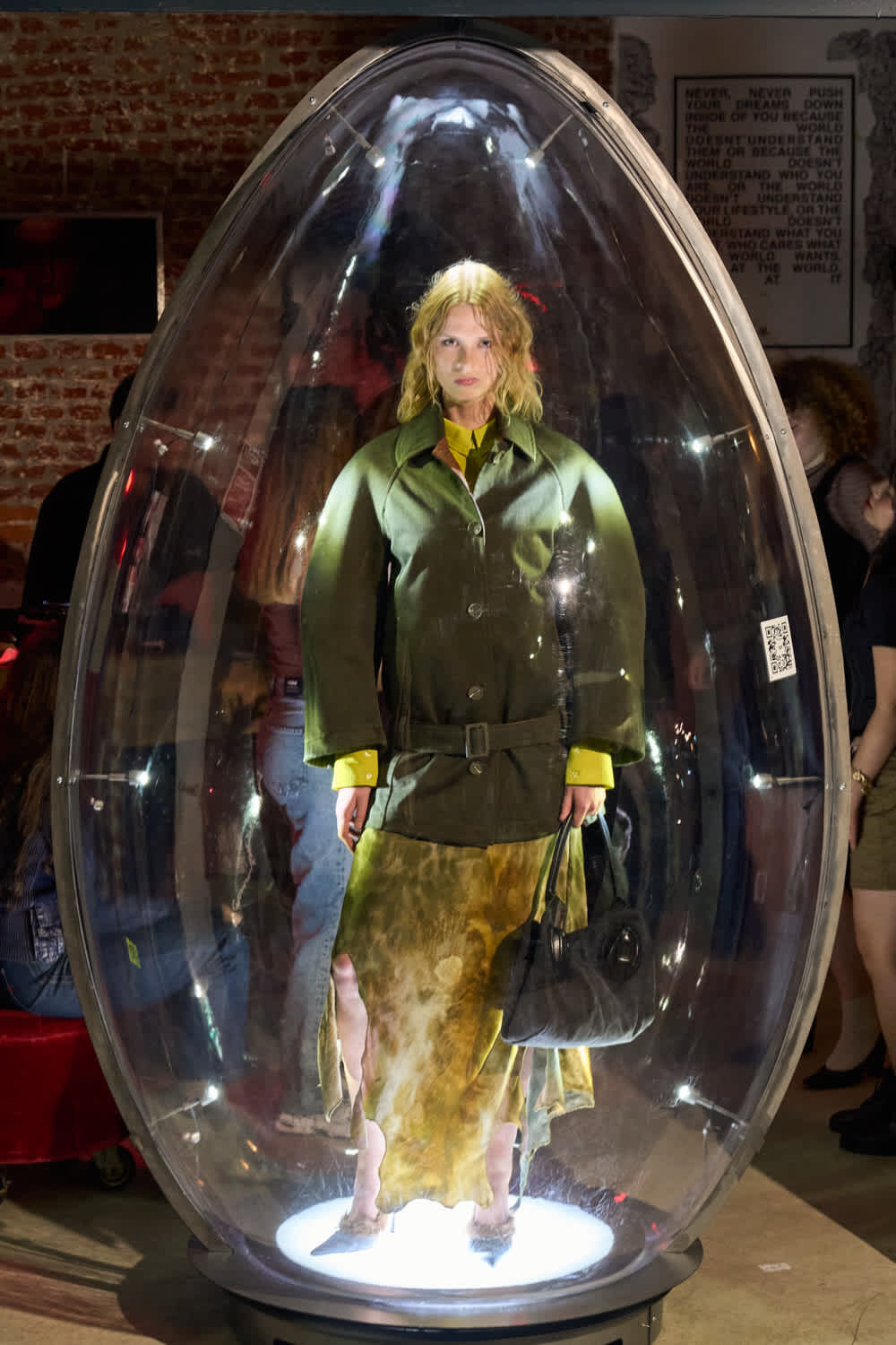 Woman in an olive-green coat and yellow cuffs stands inside a clear glass capsule on a museum floor, holding a black handbag in front of a brick wall backdrop.