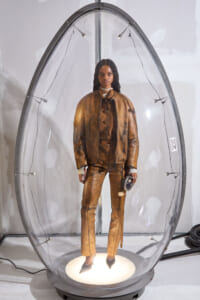 Model in a brown distressed suit inside a clear oval capsule on a lit circular base, holding a metallic helmet in a gallery setting.