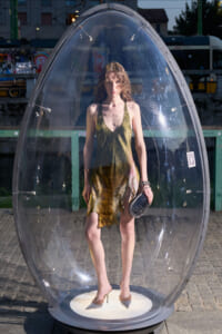Woman in a sleeveless green-brown dress stands inside a transparent, bubble-shaped capsule on a pedestal outdoors.