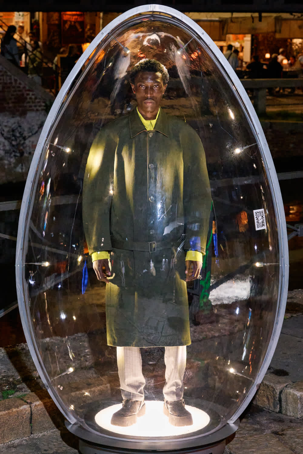 Person standing inside a clear, egg-shaped glass capsule at night, wearing a green coat and yellow shirt.
