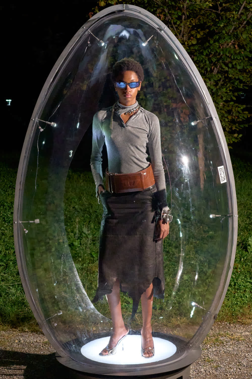 Person standing inside a clear, egg-shaped pod on a lit pedestal outdoors at night, wearing a gray long-sleeve top, black skirt, brown belt, and blue sunglasses.