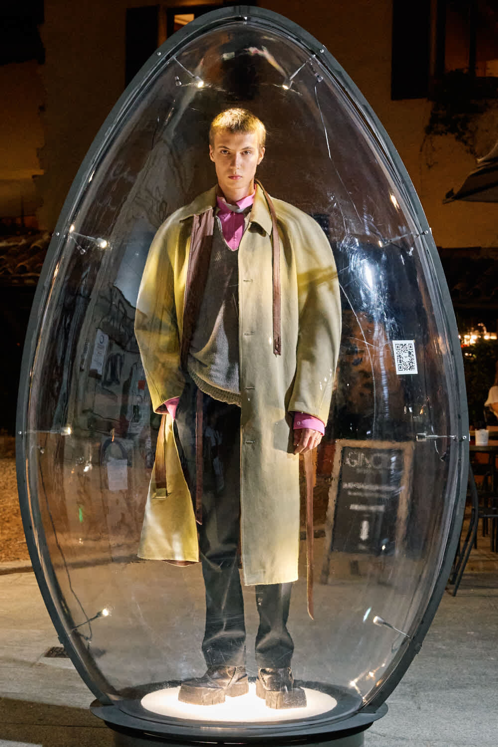 Person wearing a long beige coat over a pink shirt inside a clear, egg-shaped display capsule at night.