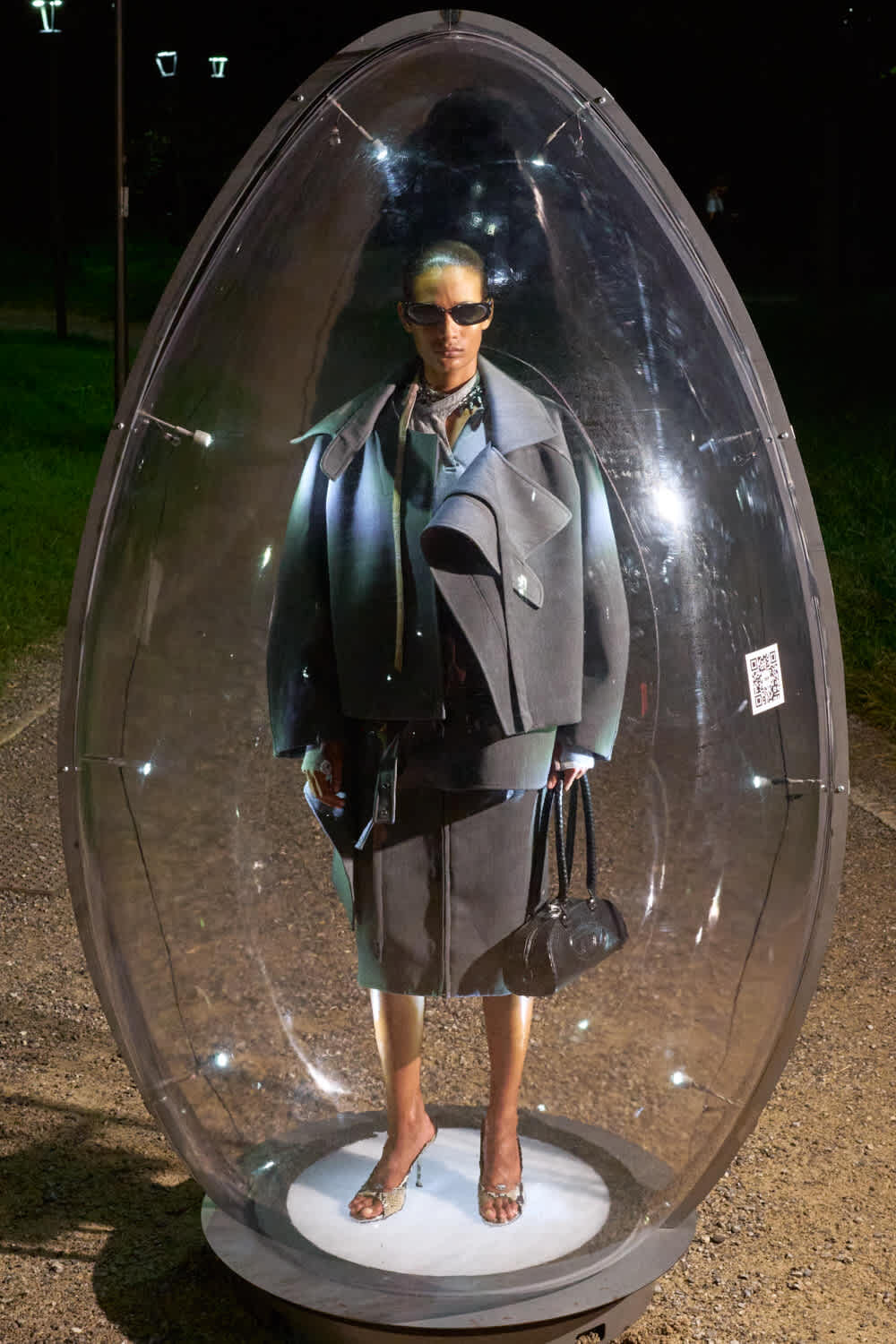Person wearing a large gray coat and sunglasses stands inside a clear glass capsule at night, holding a black handbag.