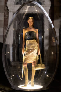 Model inside a clear glass capsule wearing a strapless satin dress, knee-high boots, and carrying a beige handbag in a gallery setting.
