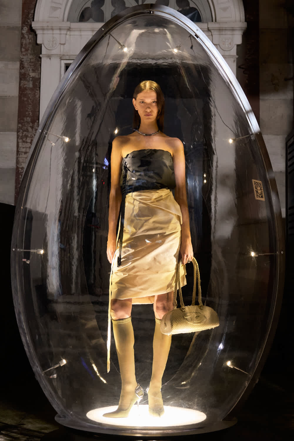 Model inside a clear glass capsule wearing a strapless satin dress, knee-high boots, and carrying a beige handbag in a gallery setting.