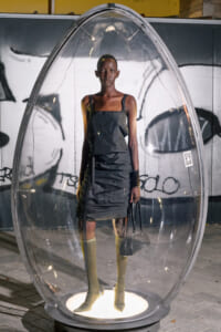 A tall dark-skinned person in a dark sleeveless dress stands inside a clear egg-shaped glass capsule on a lit pedestal, holding a black handbag with a graffiti-covered wall behind them.