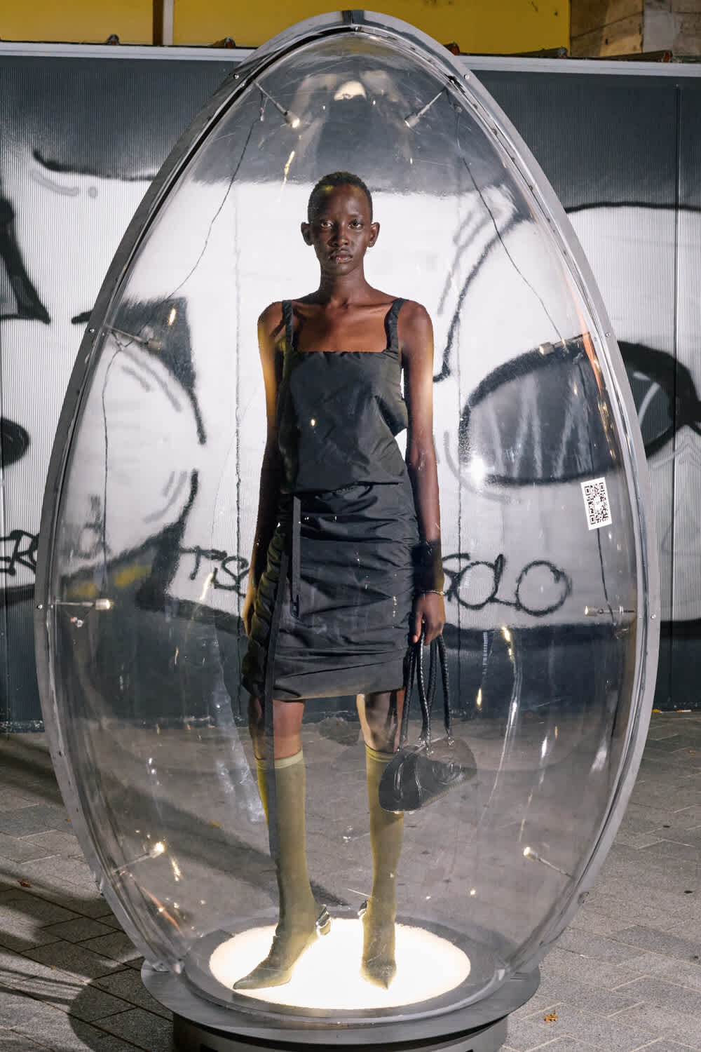 A tall dark-skinned person in a dark sleeveless dress stands inside a clear egg-shaped glass capsule on a lit pedestal, holding a black handbag with a graffiti-covered wall behind them.