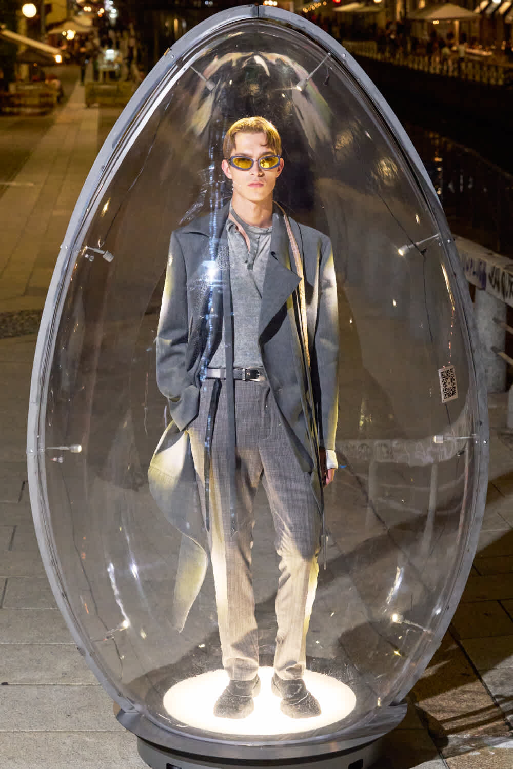 Man in a gray suit and yellow sunglasses stands inside a clear, egg-shaped glass capsule on a lit pedestal outdoors at night.