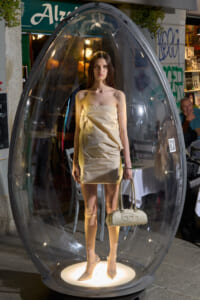 Model in a strapless metallic gold mini dress stands inside a large clear glass egg sculpture, holding a beige handbag, with a lit circular base beneath.