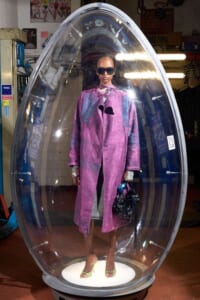 Model wearing a pink-purple coat and sunglasses inside a transparent capsule display in a workshop setting with equipment around. When read aloud: fashion display inside a clear pod.