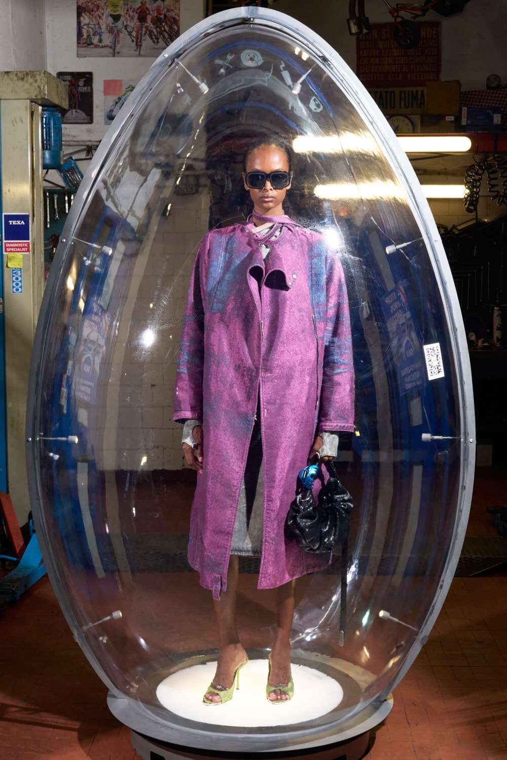 Model wearing a pink-purple coat and sunglasses inside a transparent capsule display in a workshop setting with equipment around. When read aloud: fashion display inside a clear pod.