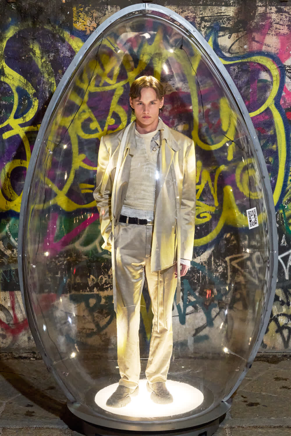 Man in a shiny gold suit stands inside a transparent glass capsule against a graffiti-covered wall, illuminated from below.