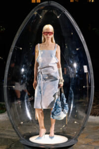 Model in a light denim dress and red sunglasses stands inside a large glass egg-shaped pod at night, holding a matching blue denim bag with beige straps.