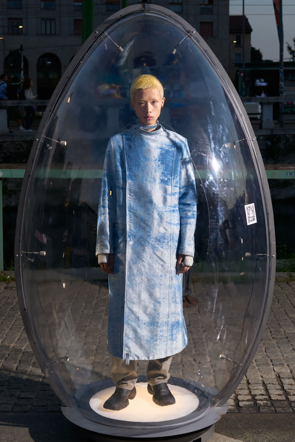 Person with bleached blond hair stands inside a transparent oval pod wearing a blue metallic coat on a cobblestone plaza.