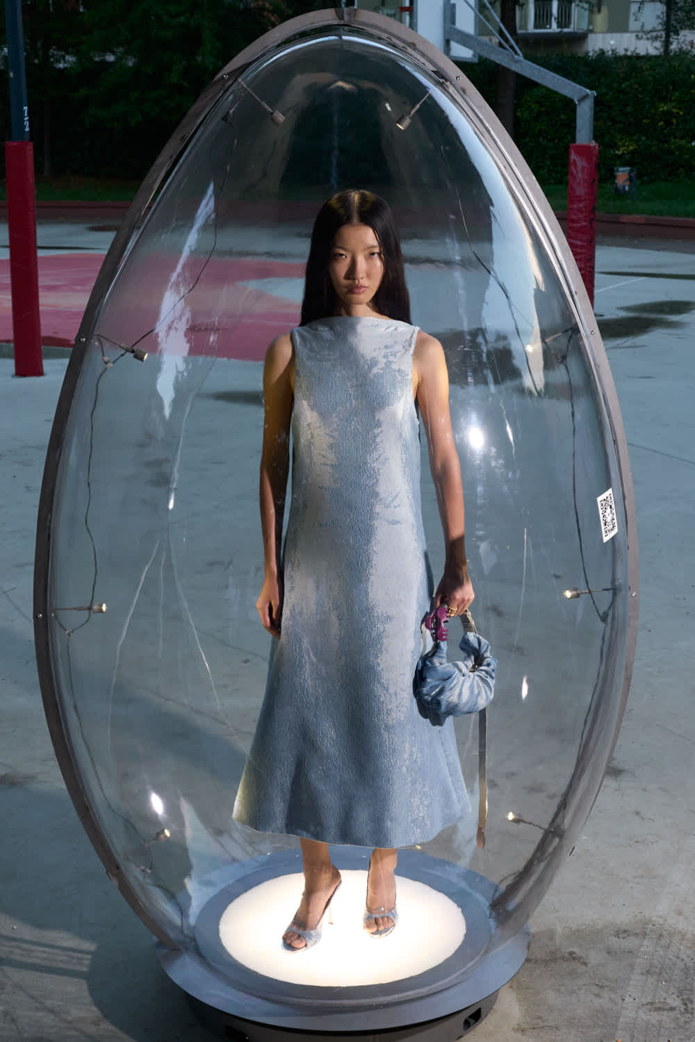 Woman in a pale blue sleeveless dress stands inside a clear glass capsule on a circular lit platform outdoors, holding a blue bag.